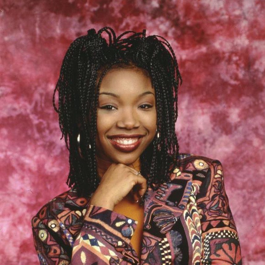A smiling woman with micro braids and hoop earrings posing against a pink background.