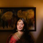 A bride in red traditional Indian clothing and gold jewelry stands with eyes closed.