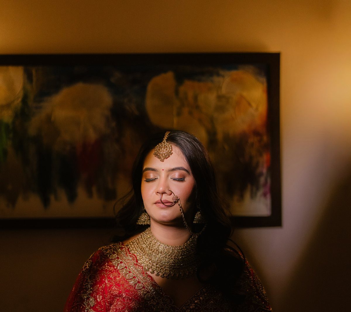A bride in red traditional Indian clothing and gold jewelry stands with eyes closed.