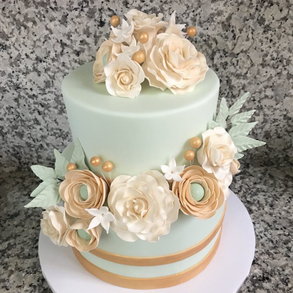 A two-tiered mint green cake adorned with cream sugar flowers and gold accents on granite.