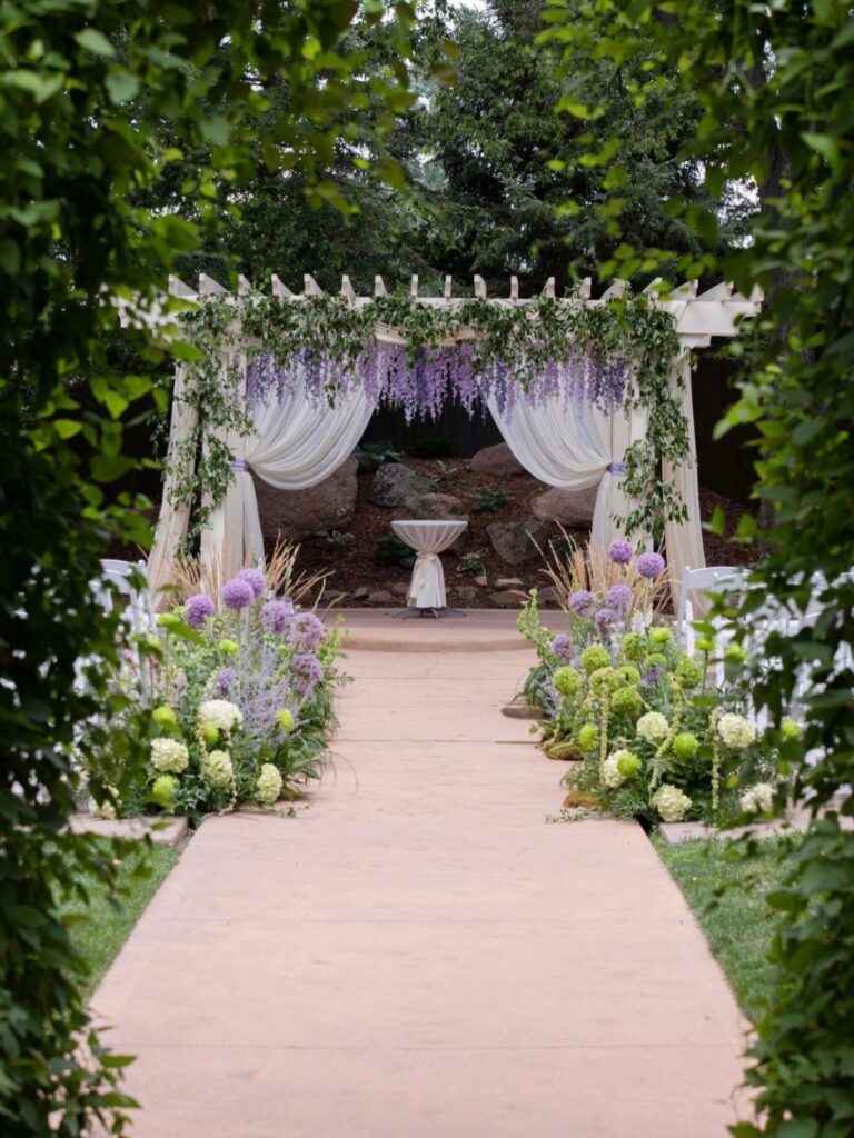 A garden wedding ceremony setup with a floral pergola and stone walkway lined with greenery.