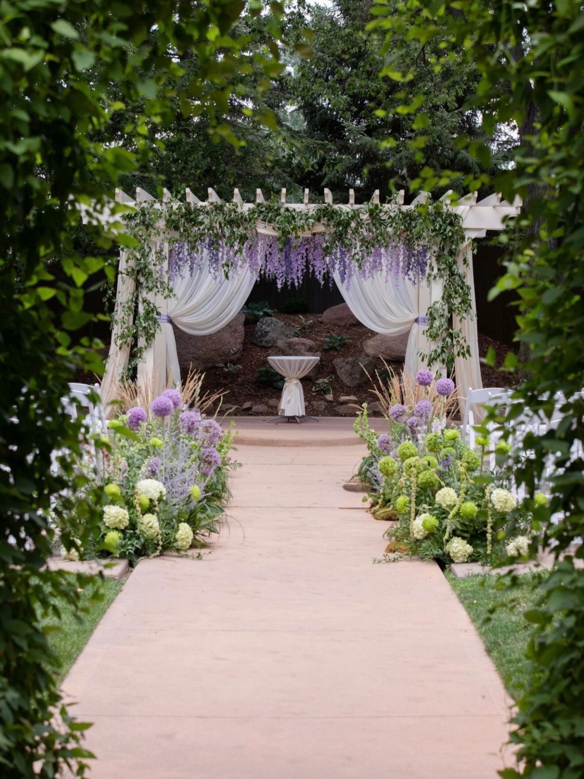A garden wedding ceremony setup with a floral pergola and stone walkway lined with greenery.