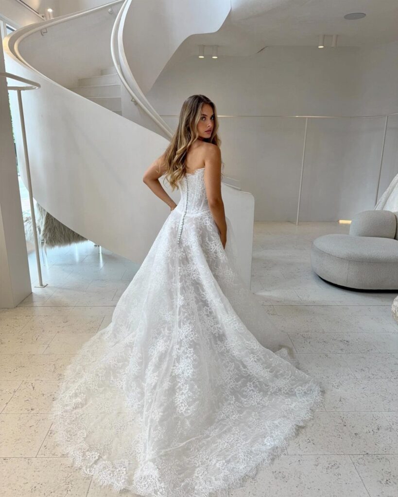 A woman in a strapless white lace wedding dress with a long train standing in a white room.