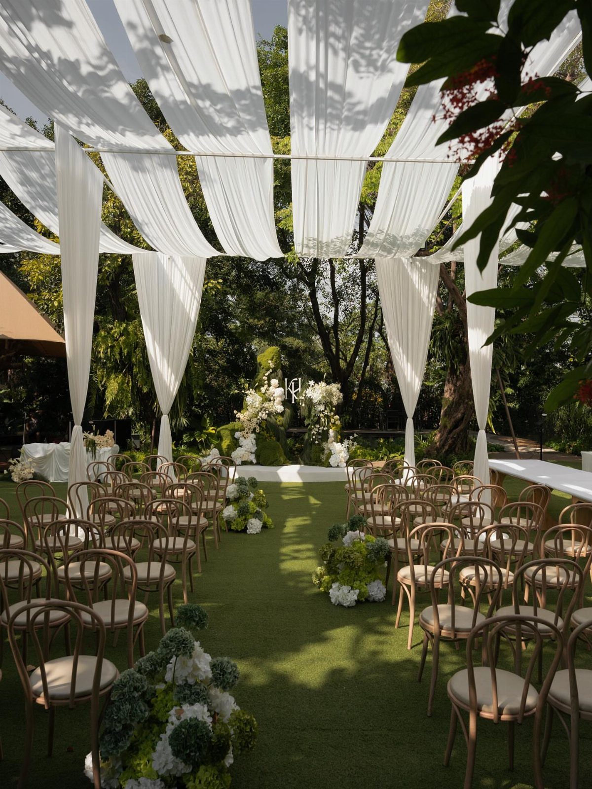 White fabric drapes hang over wooden chairs arranged for an outdoor garden wedding ceremony.