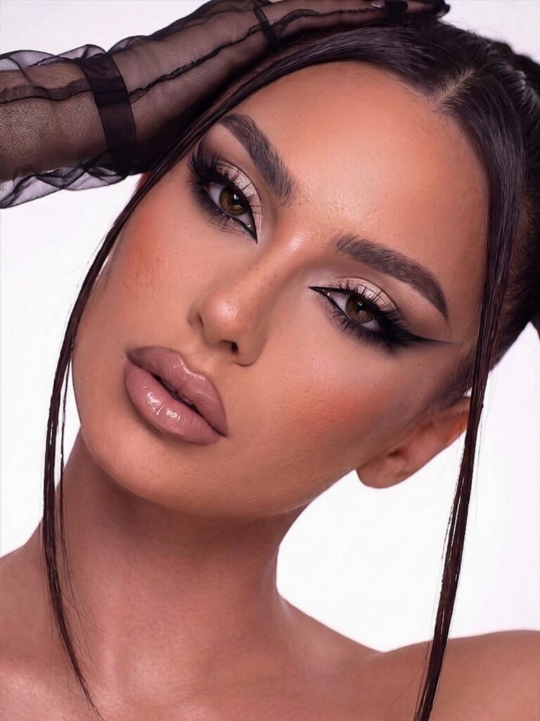 Woman with dramatic winged eyeliner, shimmery eyeshadow, and glossy lips posing against a white background.