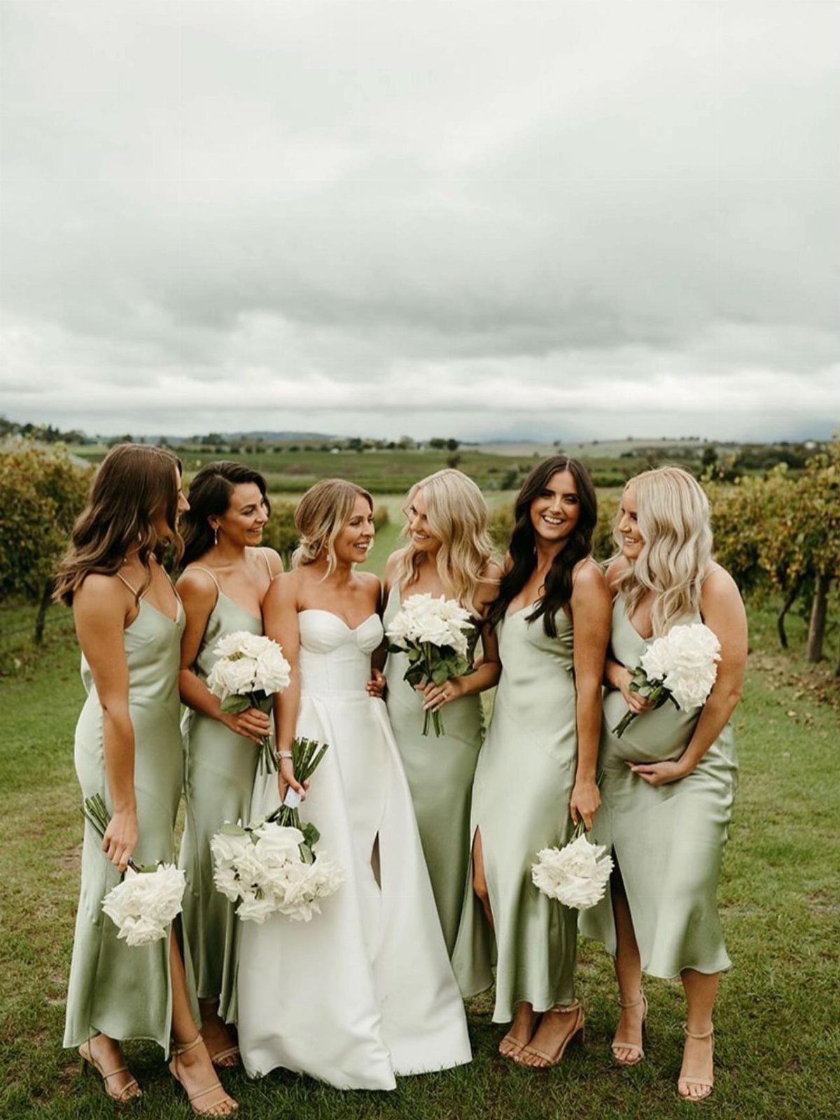 Bridesmaids in sage green dresses and a bride in white standing in a field.