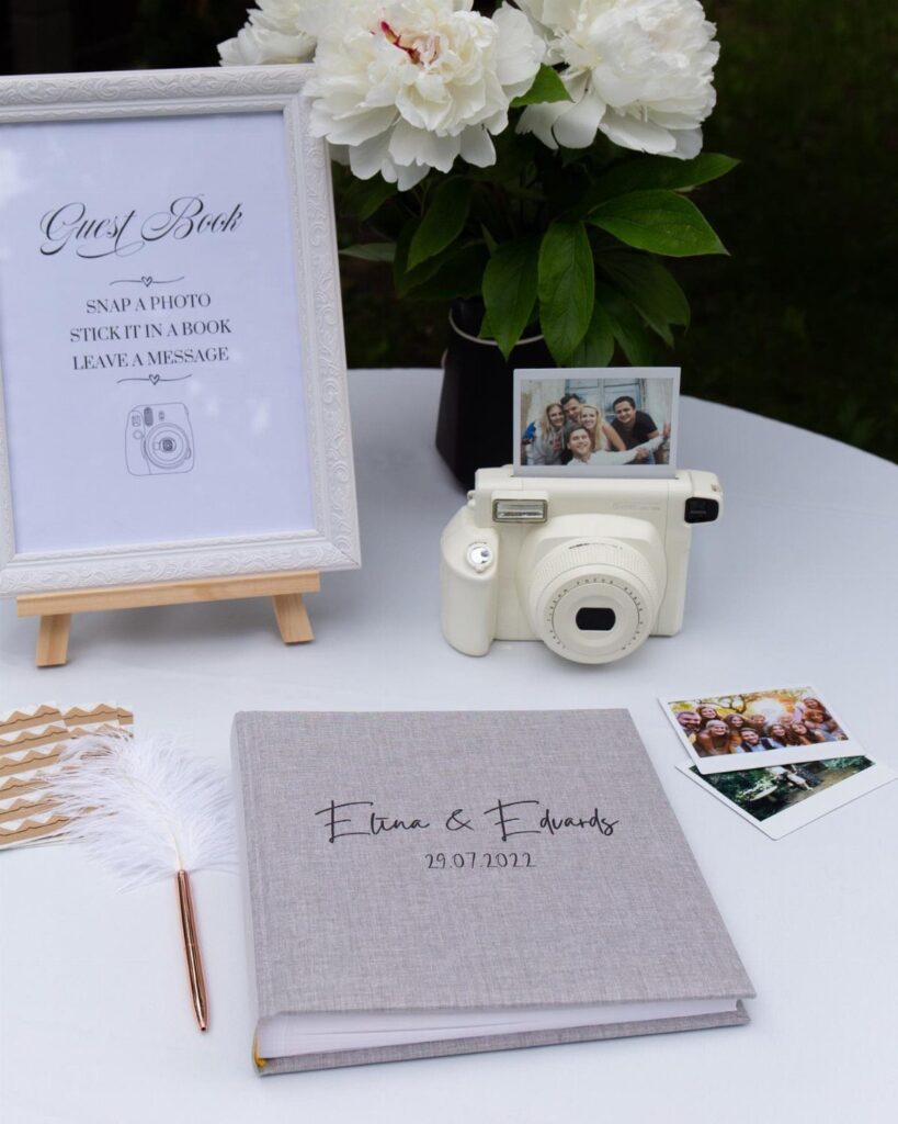 A white instant camera, a guest book, and small Polaroid photos on a white table.
