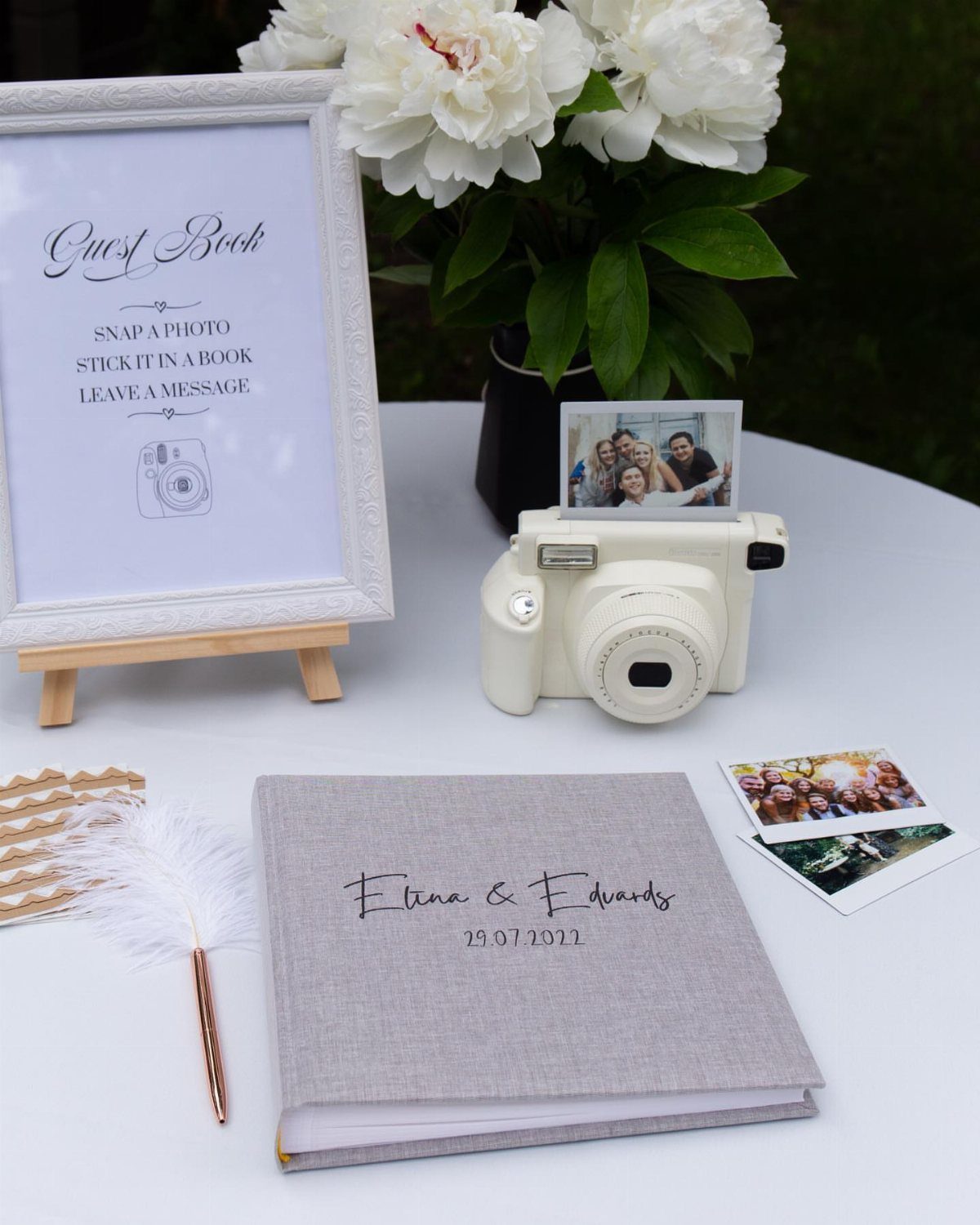 A white instant camera, a guest book, and small Polaroid photos on a white table.
