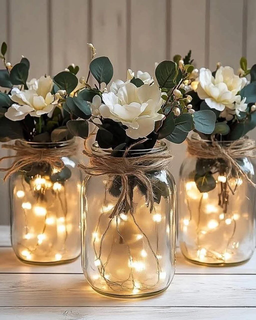 Three mason jars containing white flowers, greenery, and glowing fairy lights tied with twine.