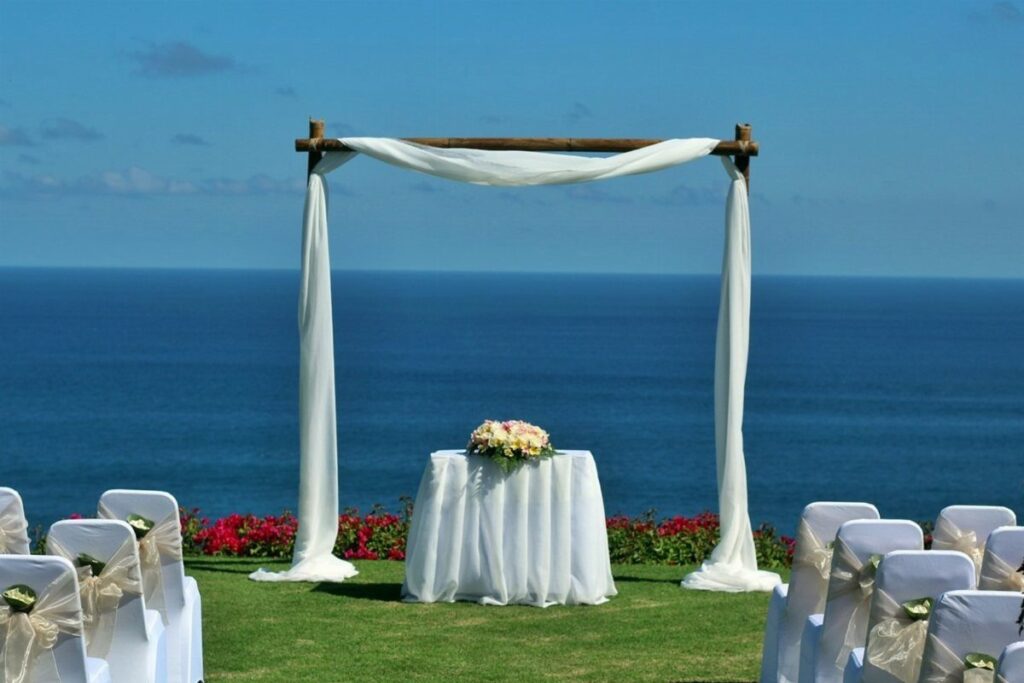 A wooden wedding arch with white fabric draped over it overlooking the blue ocean.