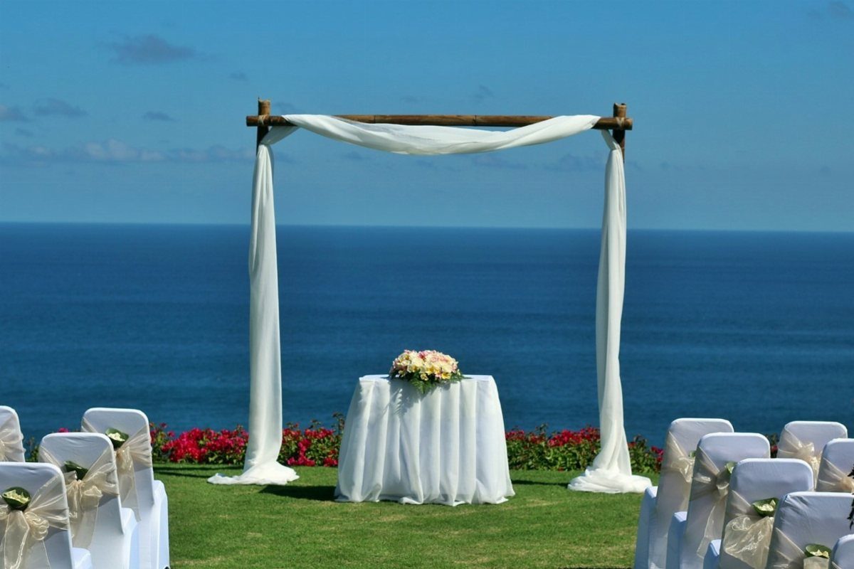A wooden wedding arch with white fabric draped over it overlooking the blue ocean.