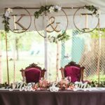Wedding sweetheart table with gold hoop backdrop, floral accents, and decorative initials under a white tent.