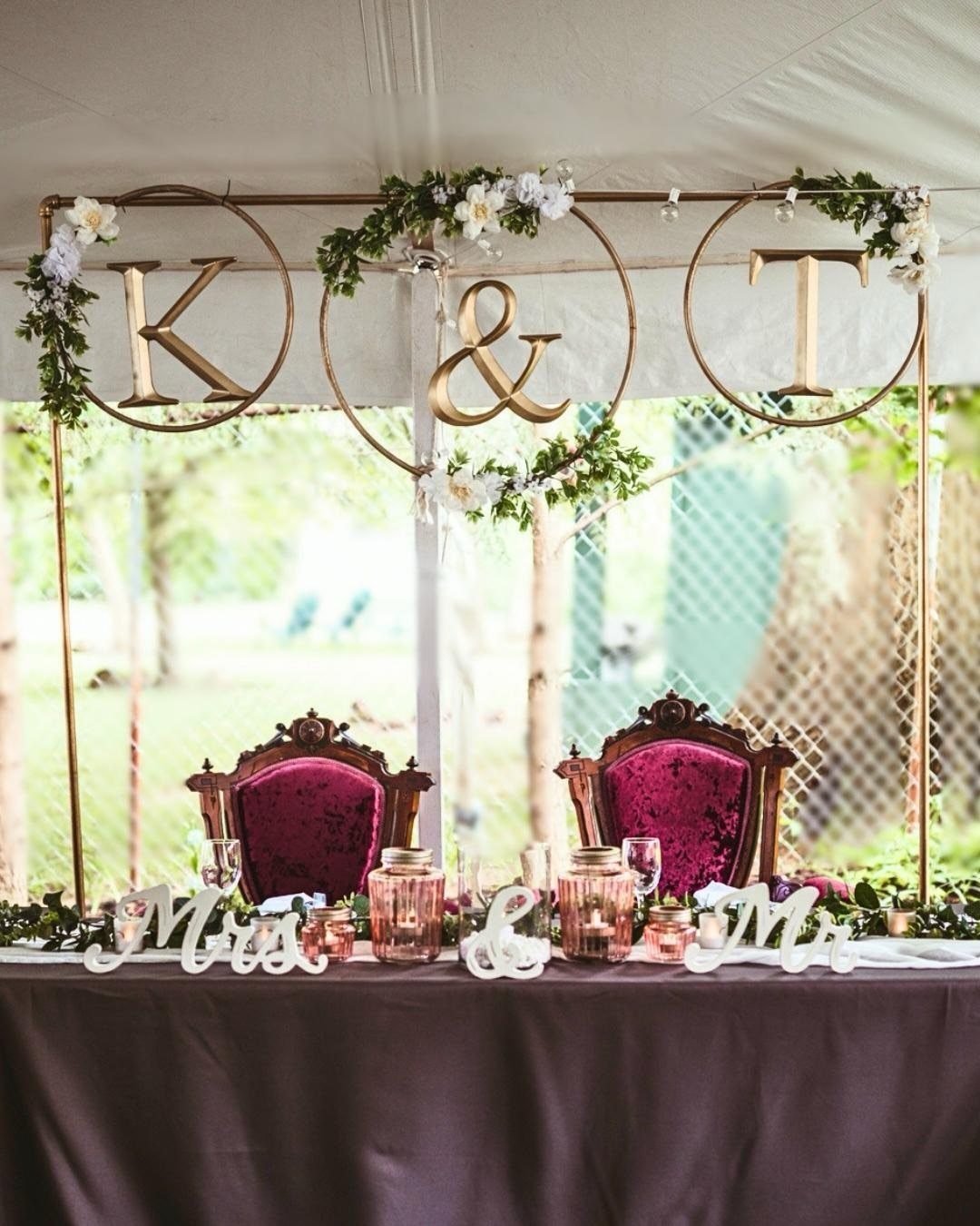 Wedding sweetheart table with gold hoop backdrop, floral accents, and decorative initials under a white tent.
