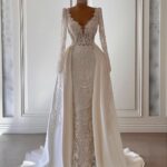 A form-fitting white lace wedding dress with long sleeves and a flowing satin overskirt.