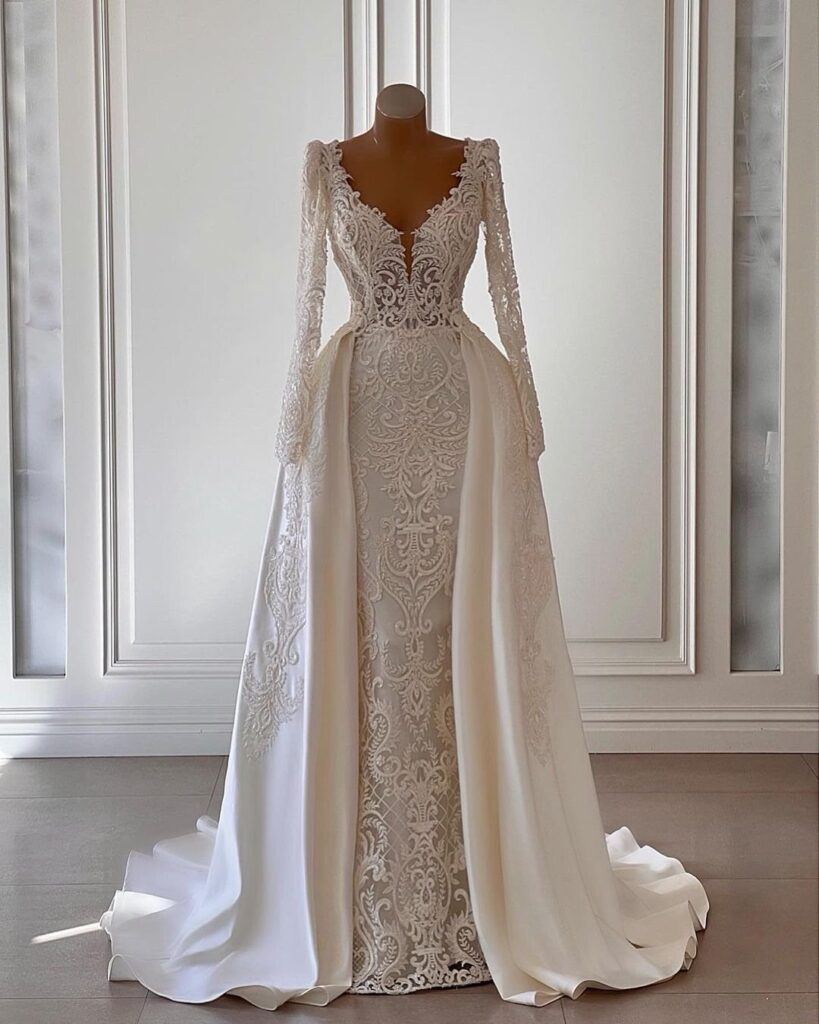 A form-fitting white lace wedding dress with long sleeves and a flowing satin overskirt.