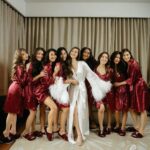 Nine women in satin robes, one in white and others in red, posing together indoors.