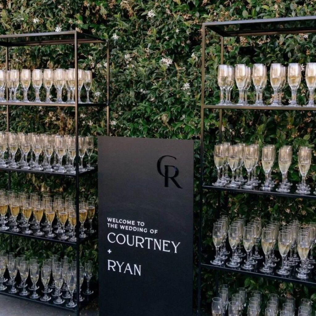 Champagne flutes arranged on black metal shelves against a green hedge with a wedding welcome sign.