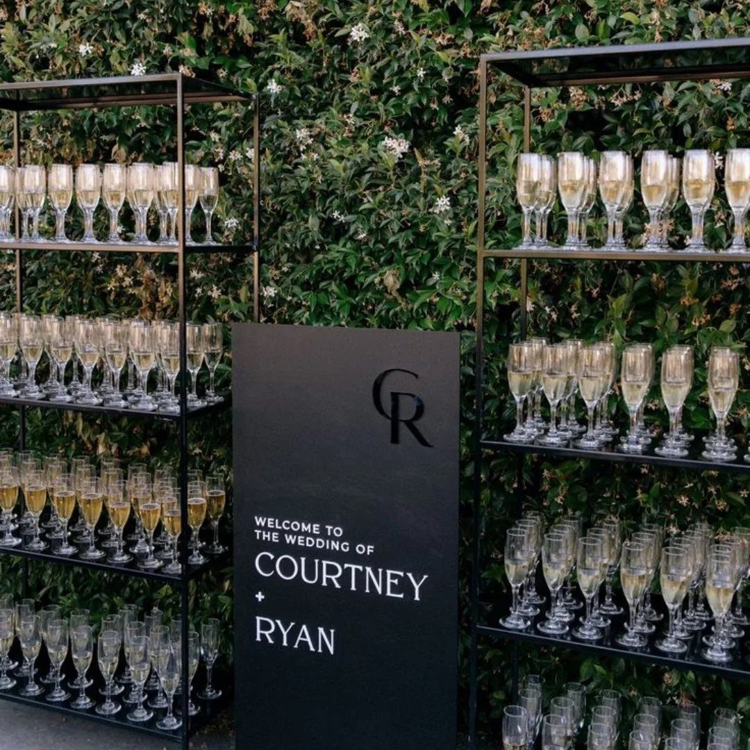 Champagne flutes arranged on black metal shelves against a green hedge with a wedding welcome sign.