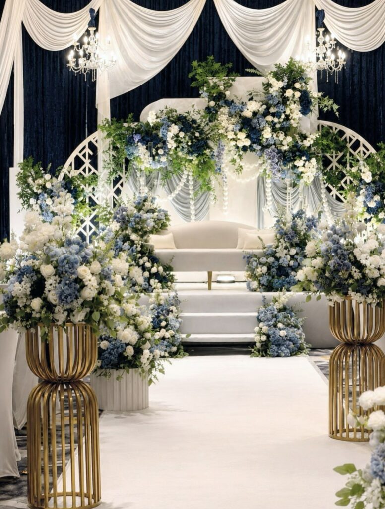 Blue and white floral arrangements decorate a wedding stage with white drapery and gold stands.