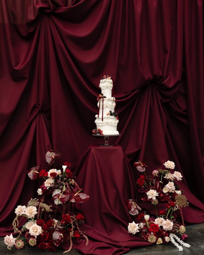 A white wedding cake on a pedestal against draped burgundy satin fabric with floral arrangements.
