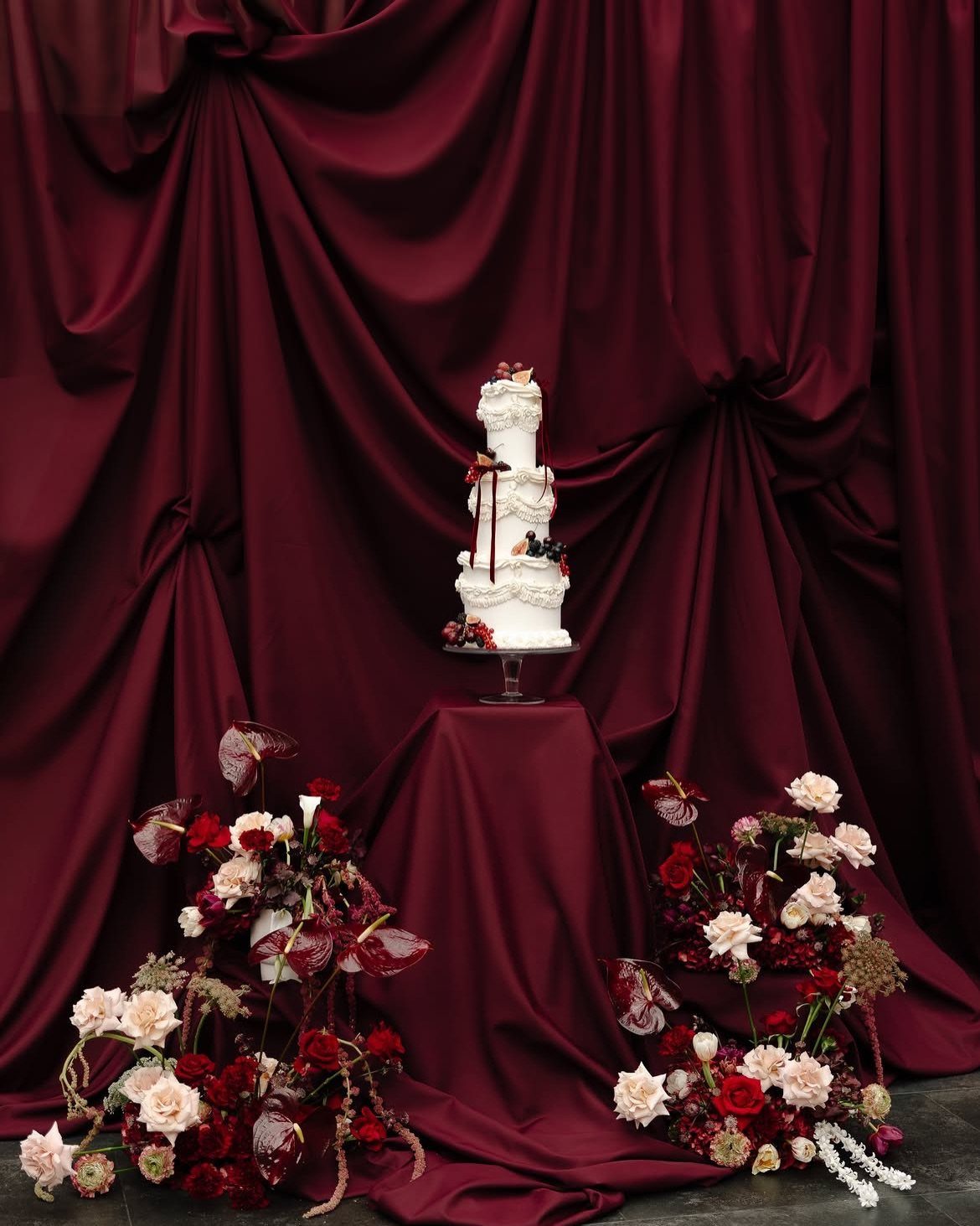 A white wedding cake on a pedestal against draped burgundy satin fabric with floral arrangements.