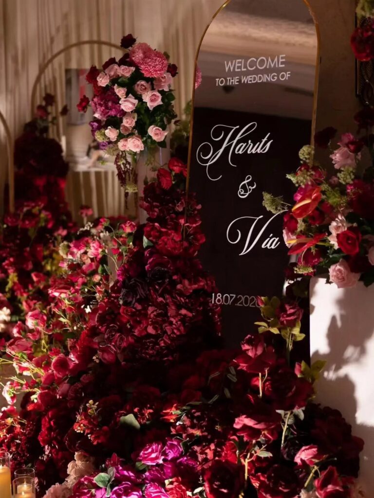 An arched black wedding welcome sign surrounded by abundant red and pink rose floral arrangements.