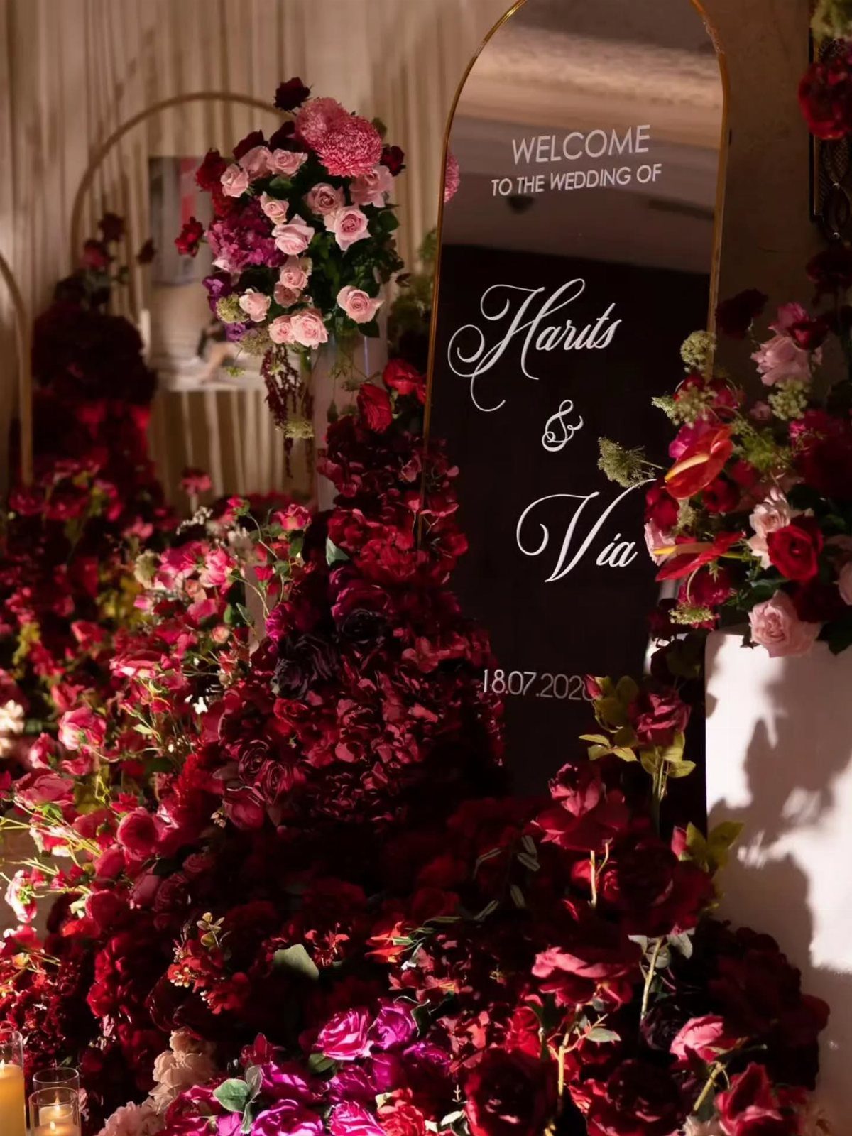 An arched black wedding welcome sign surrounded by abundant red and pink rose floral arrangements.