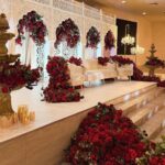 Red rose floral arrangements decorating a white wedding stage with a cream sofa and ornate backdrop.
