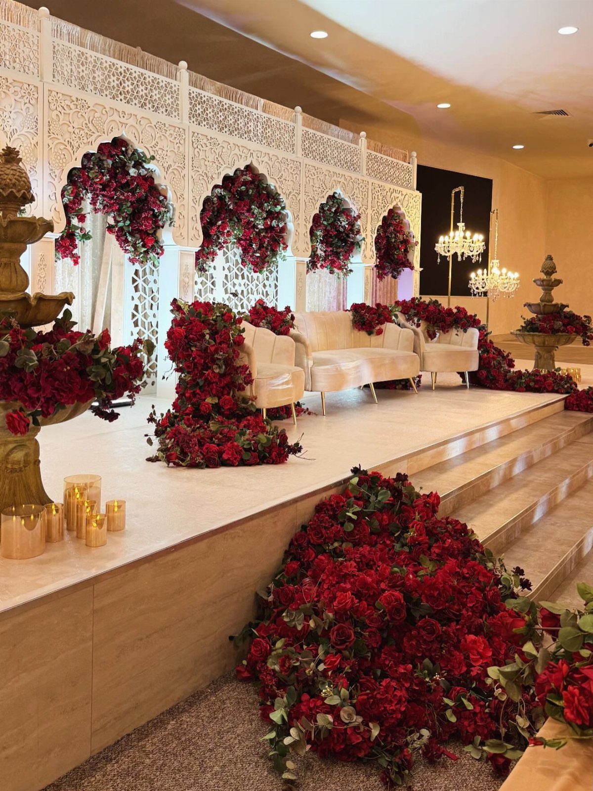Red rose floral arrangements decorating a white wedding stage with a cream sofa and ornate backdrop.