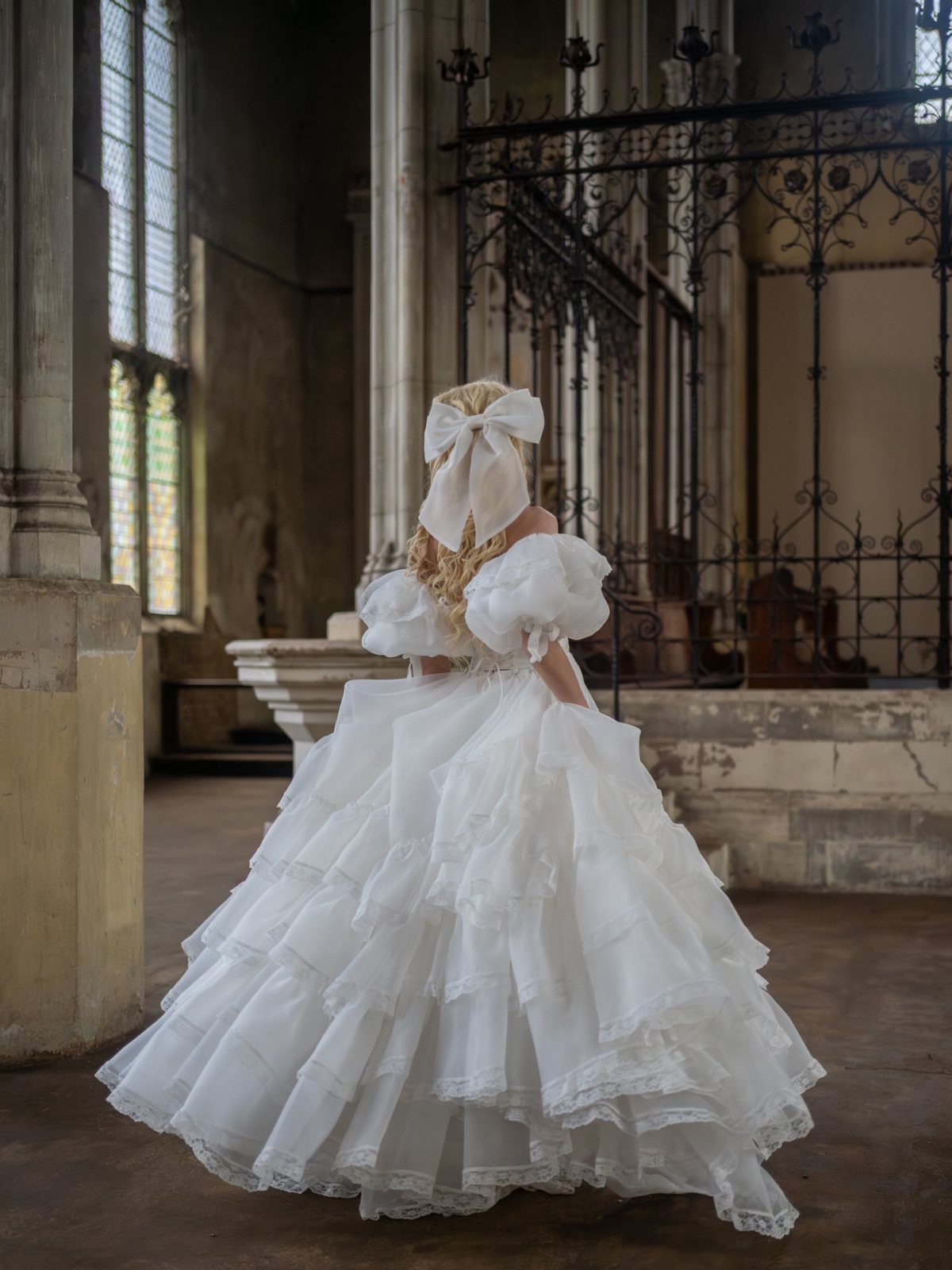 A person in a voluminous tiered white wedding dress with a large hair bow stands in a cathedral.