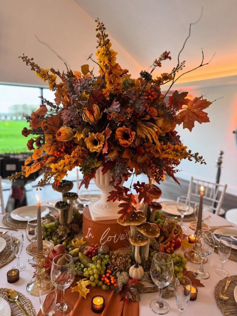 An autumnal wedding centerpiece with orange roses, maple leaves, and grapes on a decorated dining table.