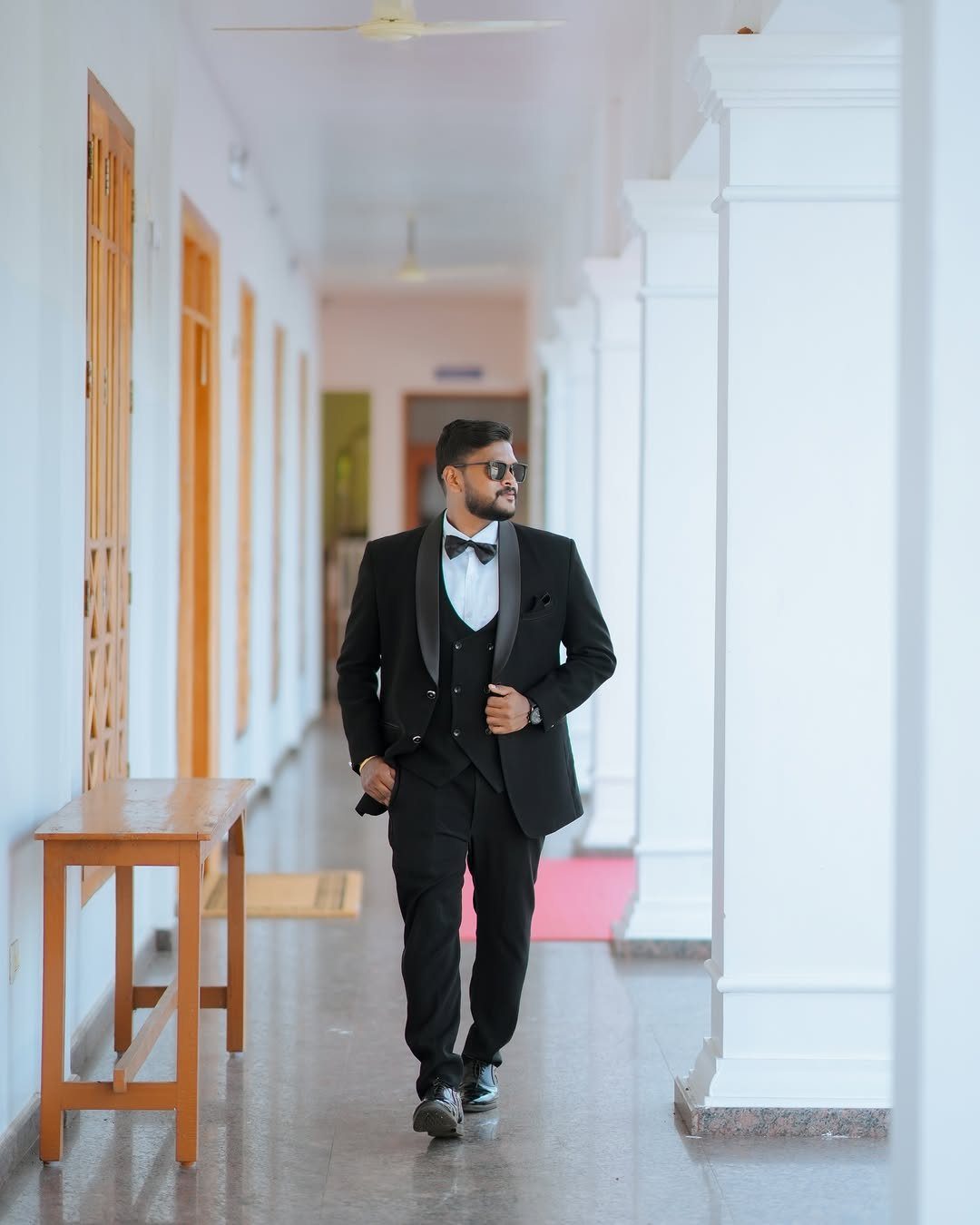 A man wearing a black tuxedo and sunglasses walks through a white hallway with columns.