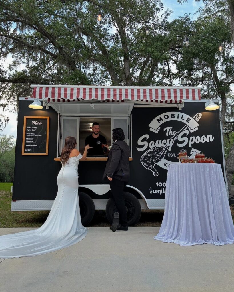 A bride and groom ordering food from a black mobile food truck outdoors.
