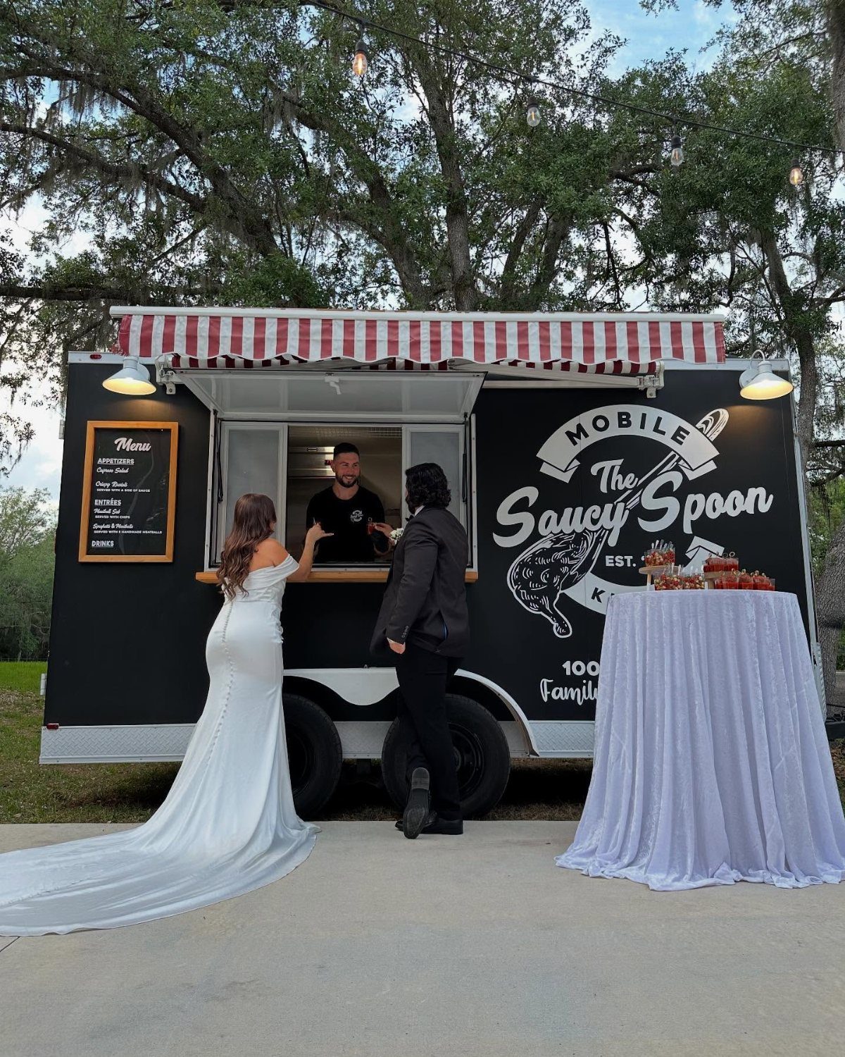 A bride and groom ordering food from a black mobile food truck outdoors.