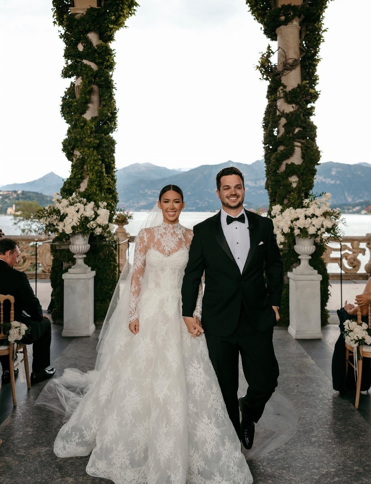 A bride in lace and a groom in a tuxedo walking together at an outdoor wedding venue.