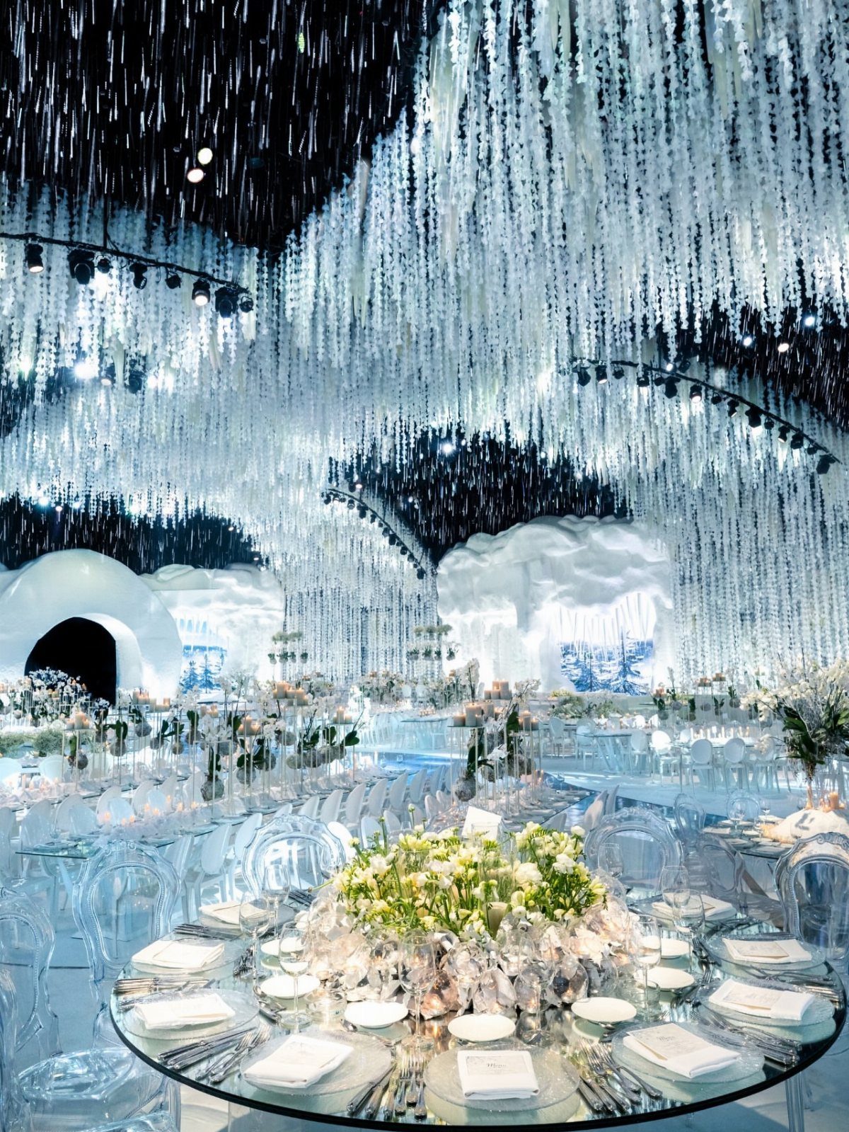 Elaborate winter wonderland wedding reception with glass tables, white flowers, and cascading ceiling decorations.