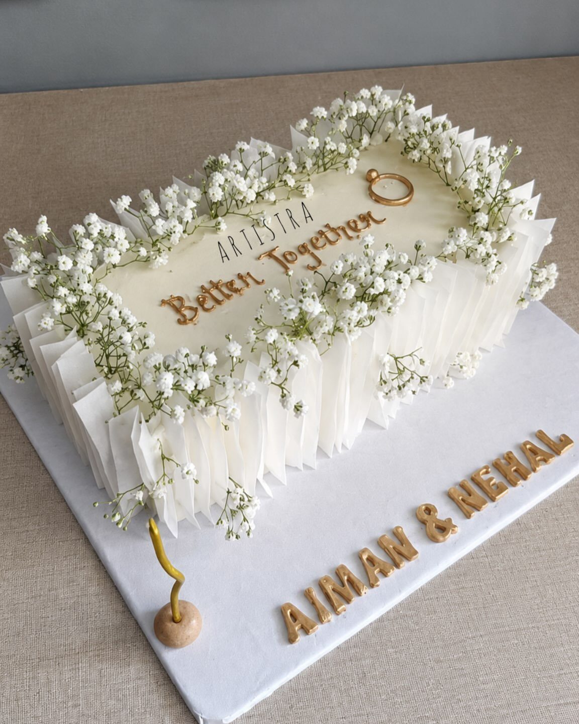 A rectangular white wedding cake decorated with baby's breath flowers and gold lettering on a white board.